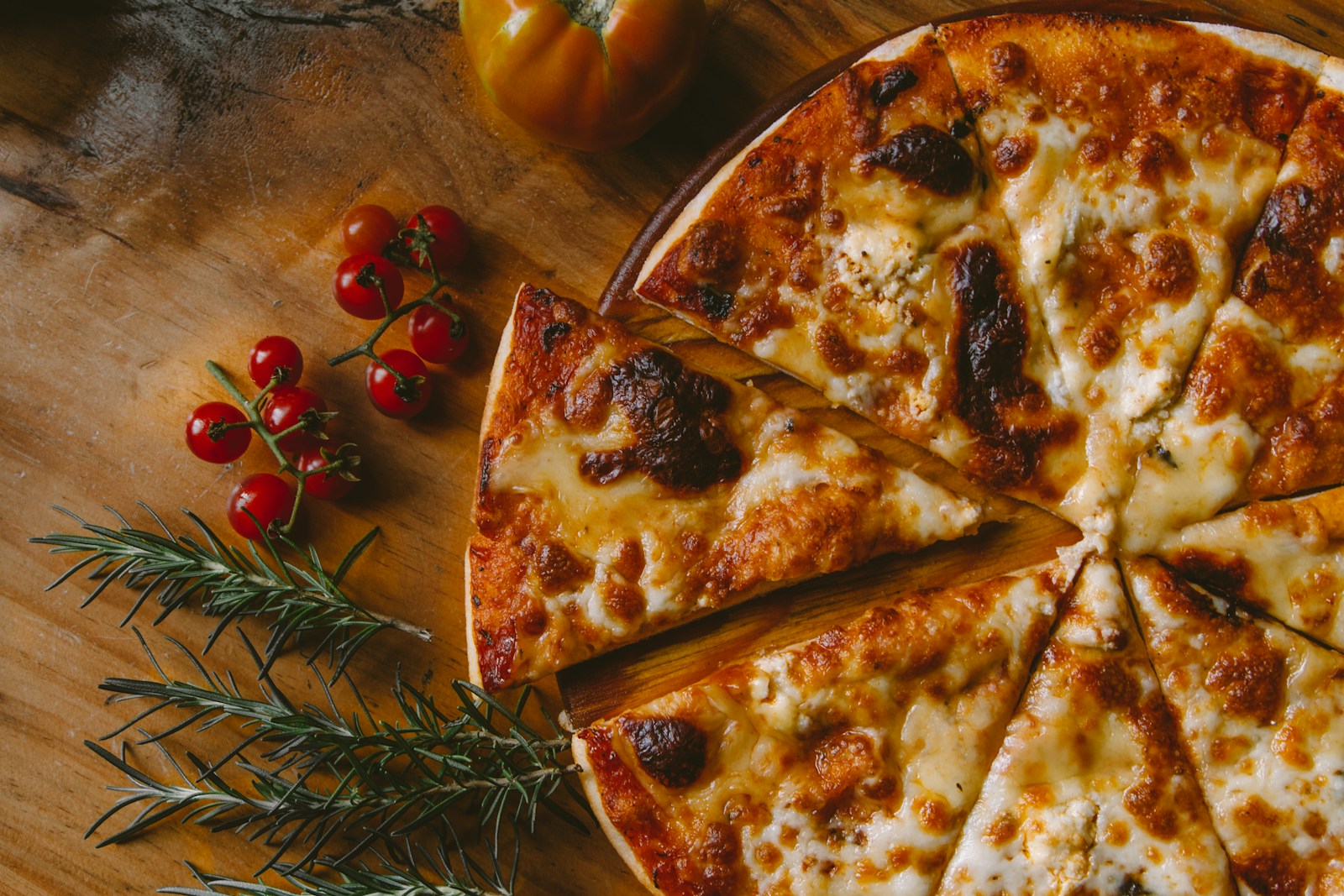 Sliced cheese pizza with tomatoes and rosemary on a wooden board.